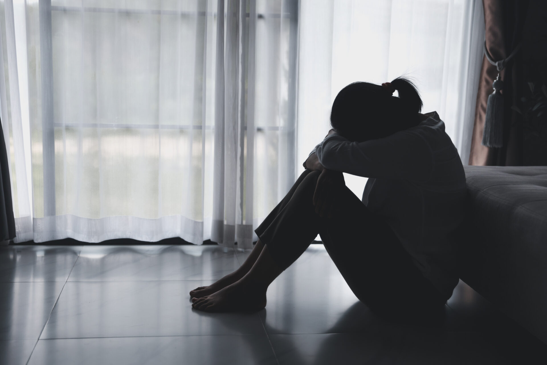 A person sits on the floor indoors with their head resting on their knees, appearing sad or distressed, silhouetted against a bright window—capturing the loneliness that can lead someone to seek a grief support group.