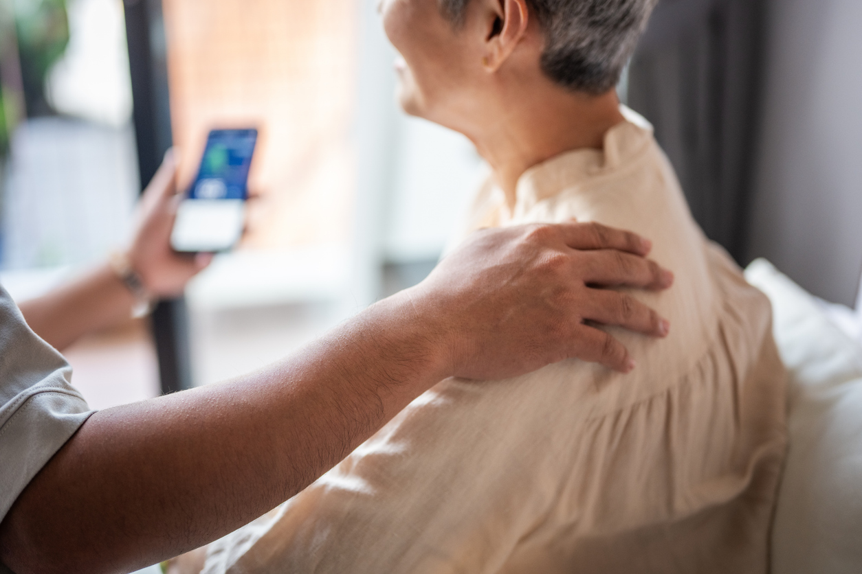 A person comforts an older adult by gently resting a hand on their shoulder, while holding a smartphone with the lifeline medical alert app open. The older adult is seated and appears to be listening.