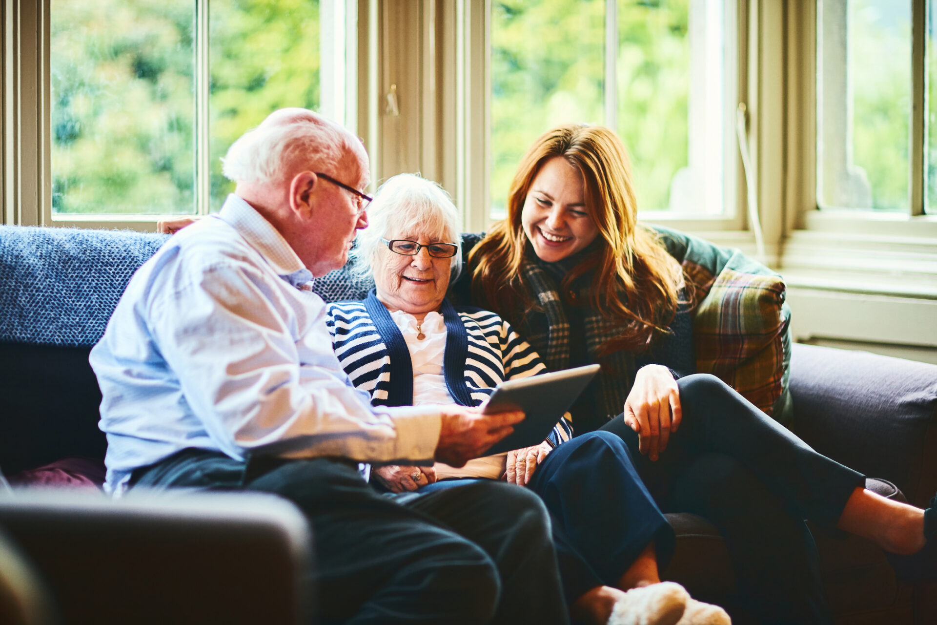 An elderly man and woman sit on a couch with a younger woman, all smiling and looking at a tablet together in a bright, cozy living room—highlighting the warmth and support of hospice care in the comfort of home.