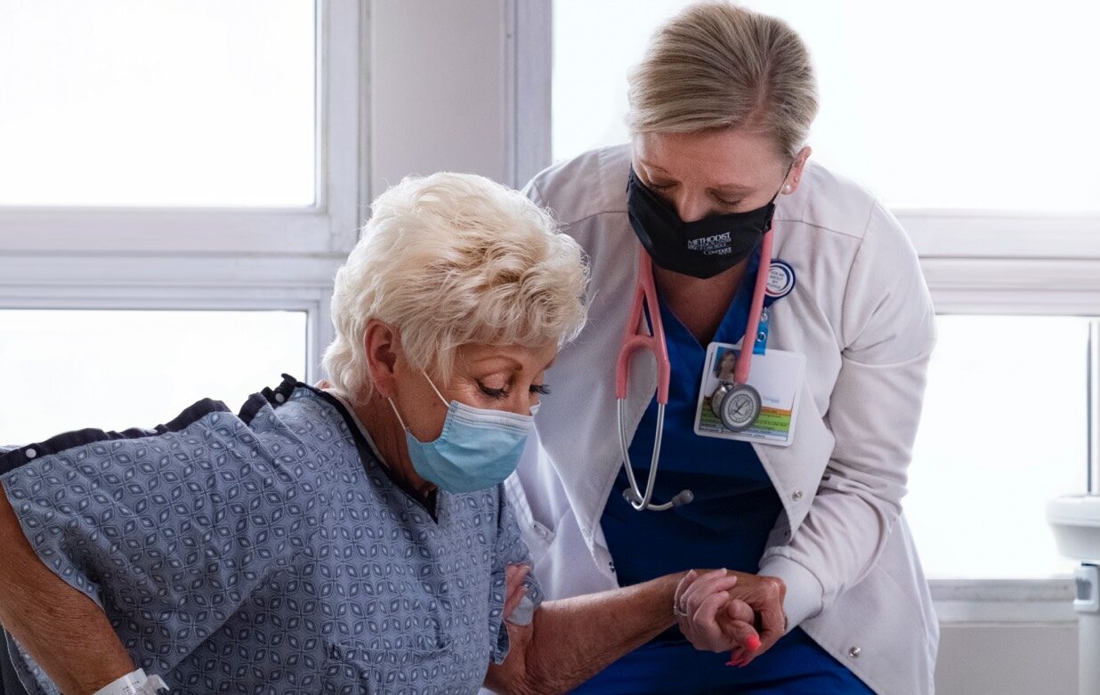 behavioral health nurse in white coat helps up an older woman in blue gown