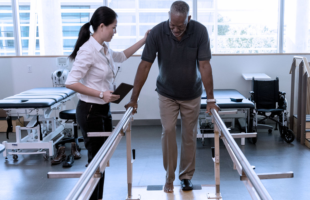 a physical therapist helps a man walk between parallel bars while he recovers from a stroke