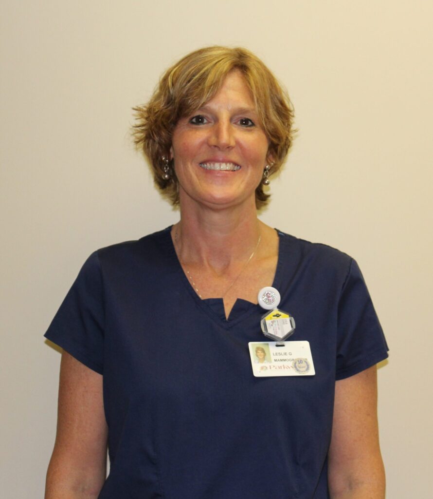 A woman with short, light brown hair is smiling and wearing navy blue scrubs. She has a name badge and pins attached to her top. The background is plain and light-colored.