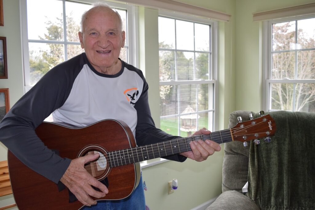 An elderly man smiles while playing an acoustic guitar, his shoulder relaxed, in a bright living room with large windows and a green recliner in the background.