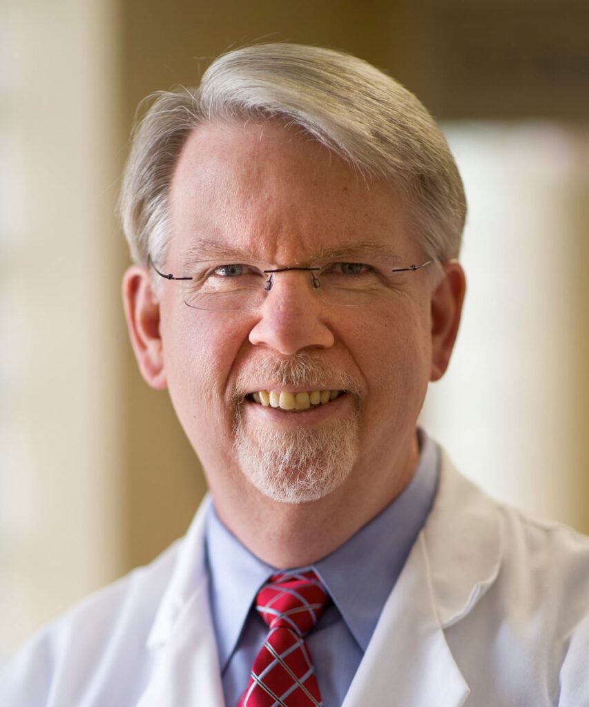 A smiling older man with gray hair, glasses, and a goatee wears a white lab coat, blue shirt, and red patterned tie—capturing the professional presence of a bariatric doctor in Knoxville. The background is softly blurred, suggesting an indoor setting.