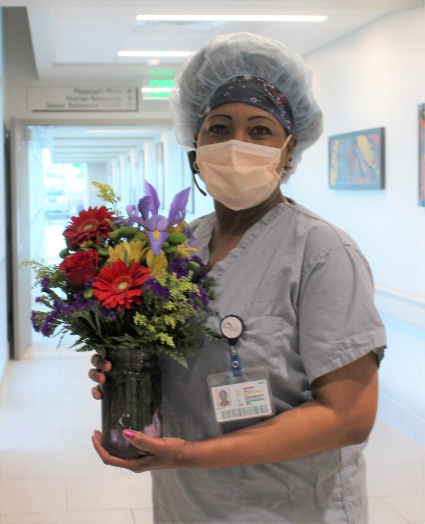 A healthcare worker wearing scrubs, a hair cap, and a face mask stands in a hospital hallway, holding a colorful bouquet of flowers.