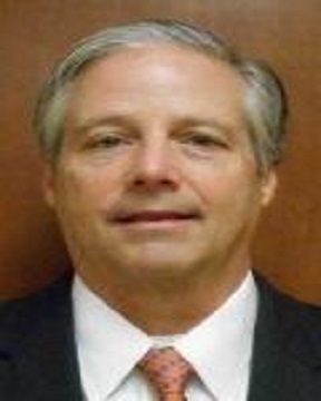 A middle-aged man with gray hair wearing a dark suit, white dress shirt, and a light-colored tie, posing against a plain brown background.