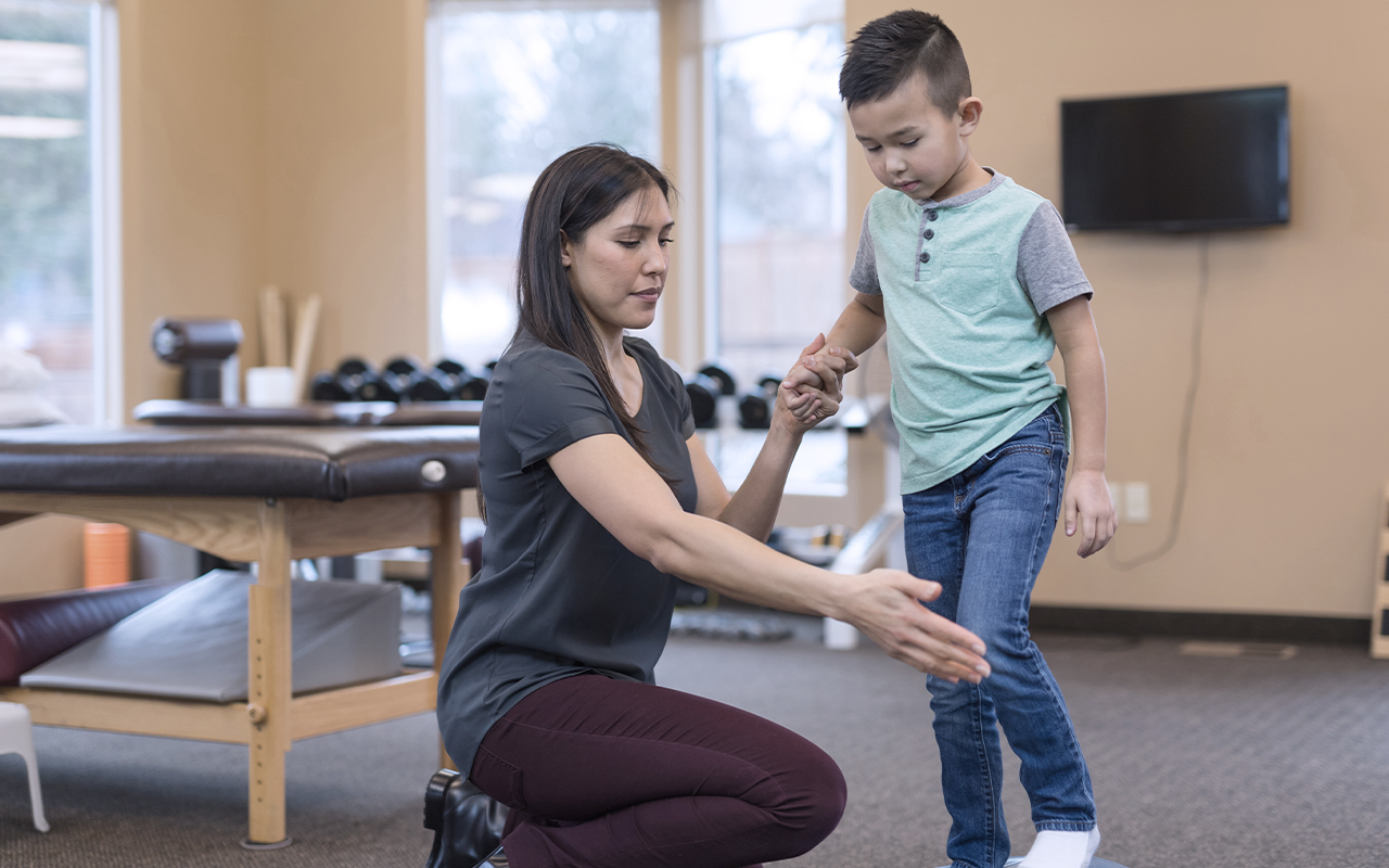 little boy balancing on one foot with help of female physical therapist