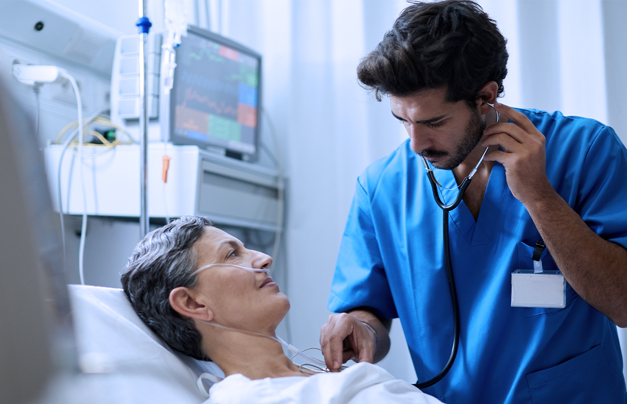 young male resident listening to the heart of a senior female patient laying in a hospital bed