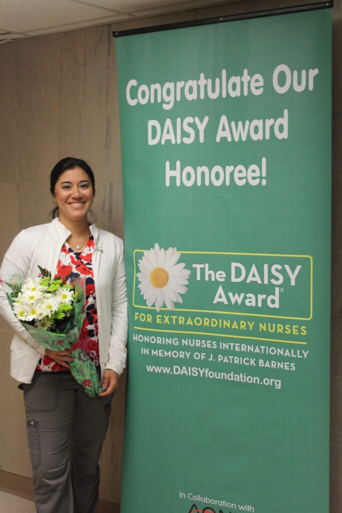 IMG_0479 A smiling nurse named Mel holds a bouquet of flowers beside a banner that reads, Congratulations Our DAISY Award Honoree! The DAISY Award for Extraordinary Nurses.