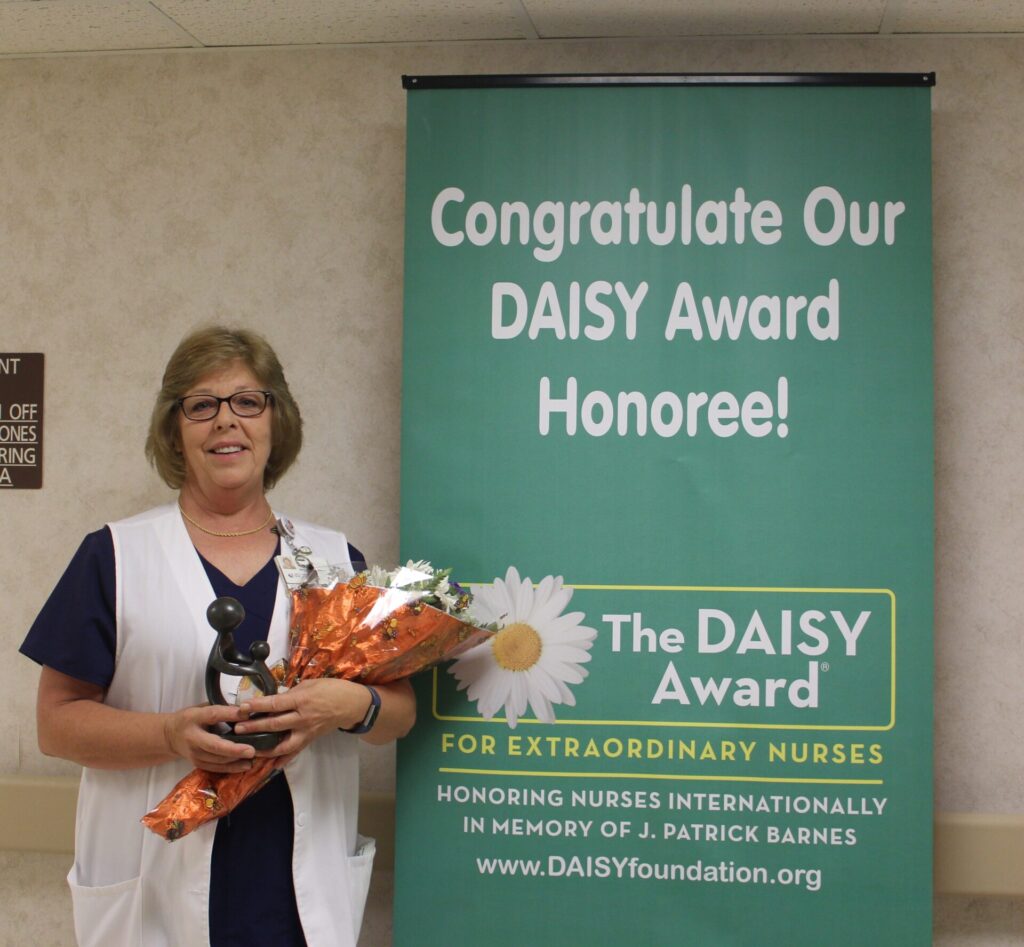 Nurse Jamie, in scrubs and a white vest, holds a bouquet and a trophy, standing next to a banner that reads Congratulate Our DAISY Award Honoree! The DAISY Award for Extraordinary Nurses.