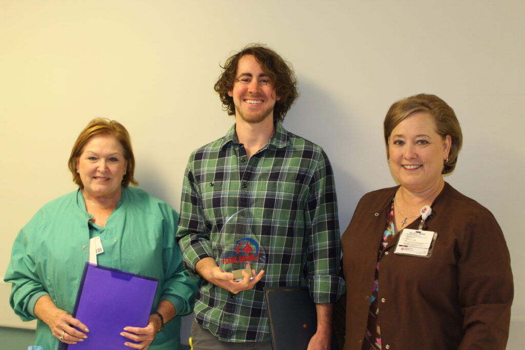 Three adults stand side by side smiling at the camera. Ian, in the center, holds a clear award trophy, while the two women on either side hold folders. They are standing against a plain background.