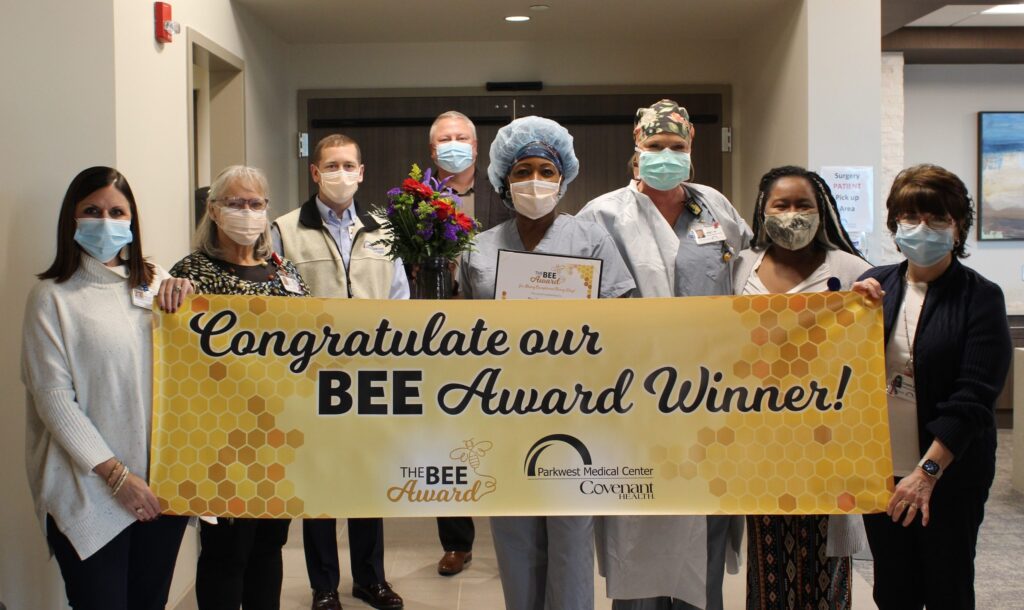 A group of masked people stands in a hallway holding a yellow banner that reads “Congratulations our BEE Award Winner!” One person in scrubs holds flowers, a certificate, and the award.