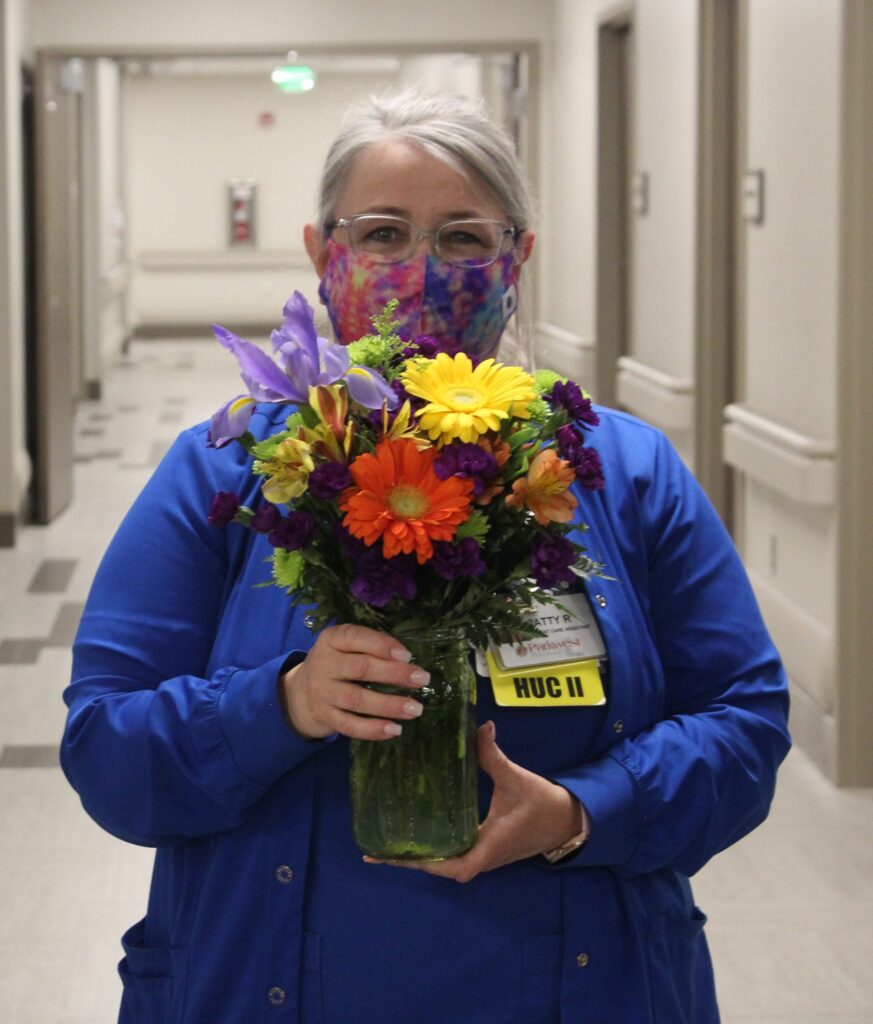 A woman wearing a colorful face mask and blue scrubs holds a bright bouquet of mixed flowers in a glass vase while standing in a hallway. She has gray hair and an ID badge clipped to her chest.