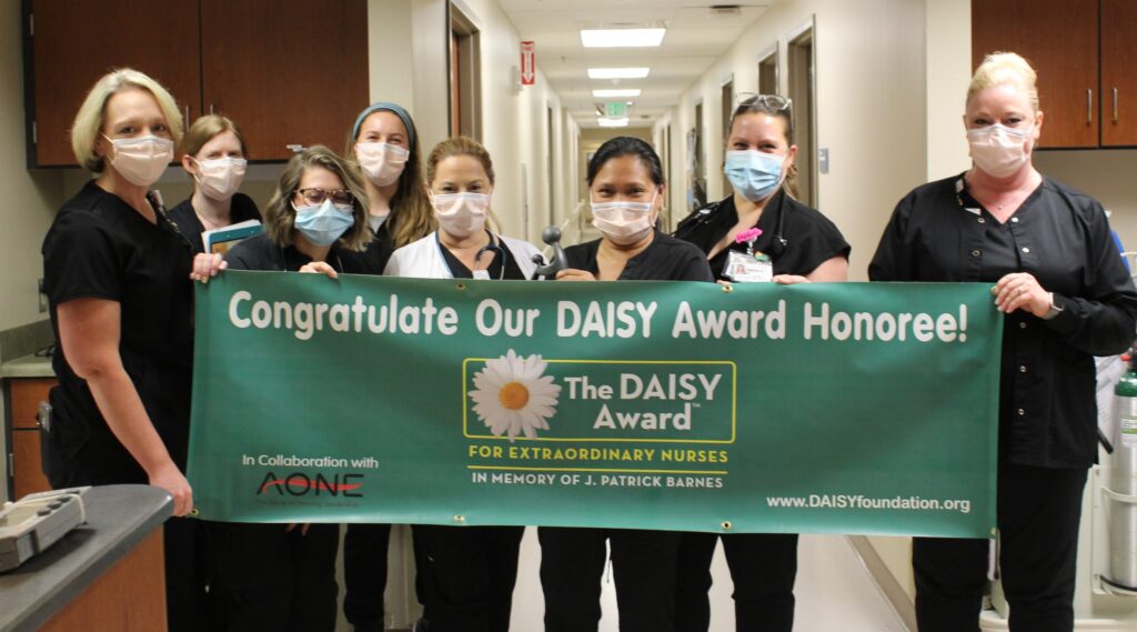 Eight nurses wearing scrubs and face masks stand in a hallway holding a green banner that reads, “Congratulate Our DAISY Award Honoree! The DAISY Award for Extraordinary Nurses.”.