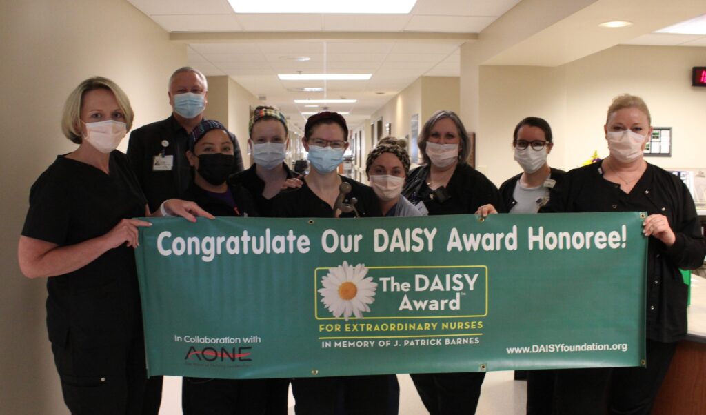 A group of nurses wearing masks stand in a hallway, holding a green banner that reads, Congratulate Our DAISY Award Honoree! The DAISY Award for Extraordinary Nurses.