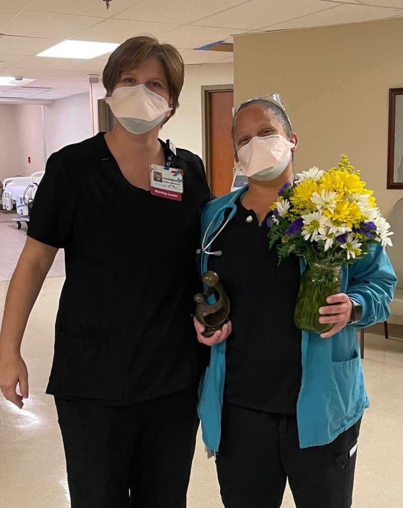 Two healthcare workers wearing masks stand together in a hallway. One holds a bouquet of yellow, white, and purple flowers and a small award. Both are smiling behind their masks. Hospital beds and equipment are in the background.