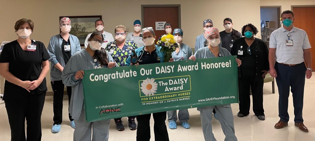 A group of healthcare workers in scrubs and masks stand indoors, holding a green banner that reads, Congratulations Our DAISY Award Honoree! The DAISY Award for Extraordinary Nurses. One person holds flowers.