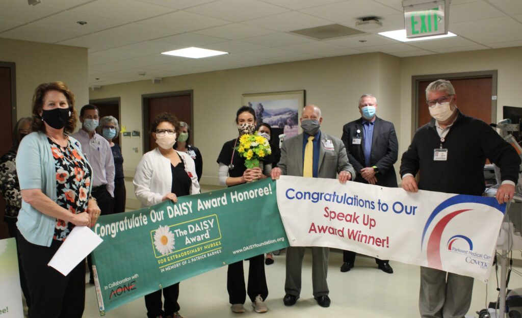 A group of healthcare workers wearing masks stand in a hospital hallway holding banners celebrating award winners. One woman holds a bouquet of flowers, and others smile for the photo.