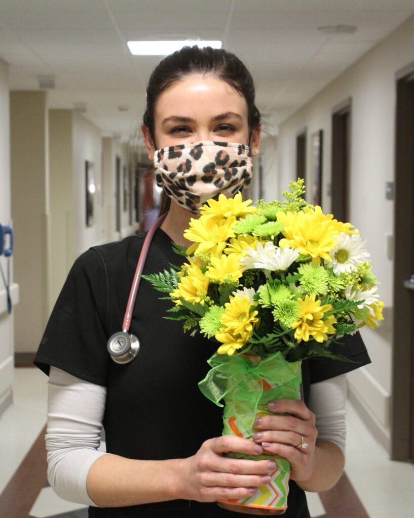 A healthcare worker wearing a leopard print mask and black scrubs holds a bouquet of yellow and white flowers while standing in a hospital hallway.