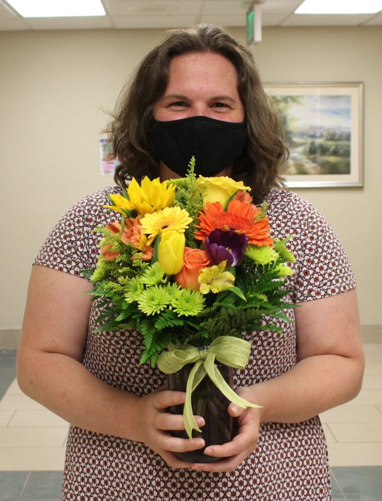 A person wearing a black face mask and patterned dress holds a bouquet of colorful flowers, including yellow, orange, and purple blooms, tied with a green ribbon, standing indoors.