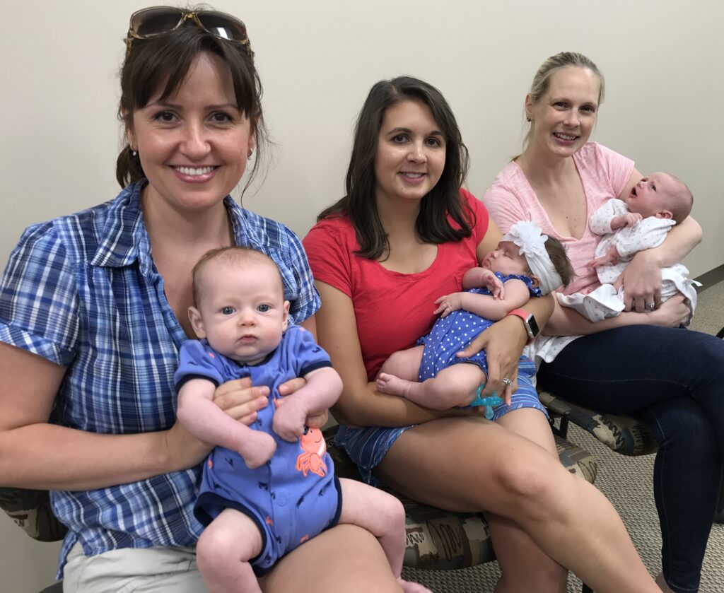 Three women sit on chairs indoors, each holding a baby. All are smiling at the camera. The babies wear colorful outfits, and one baby has a headband. The atmosphere is warm and casual.