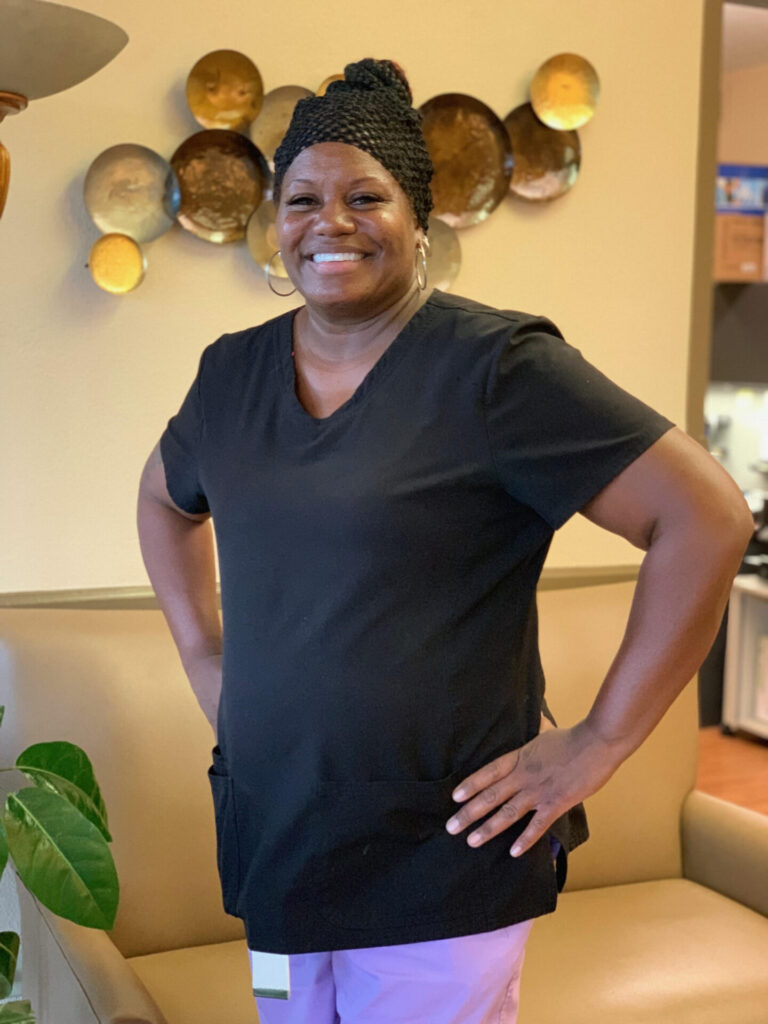 A woman wearing black scrubs and pink pants stands smiling with her hands on her hips in a warmly lit room, with decorative plates on the wall behind her.