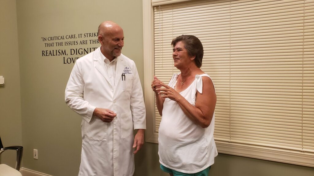 Swisher and Barnhill 3 Dr. Barnhill, in a white coat, smiles and talks with a woman in a sleeveless shirt who gestures with her hands, in a medical office with beige walls and closed blinds.