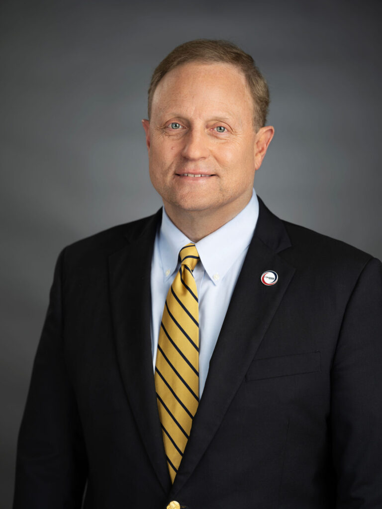 A man in a dark suit, white shirt, and yellow striped tie stands against a gray background. He has short, light brown hair and wears a circular pin on his lapel, perhaps representing what is Covenant Health. He is smiling slightly and looking at the camera.