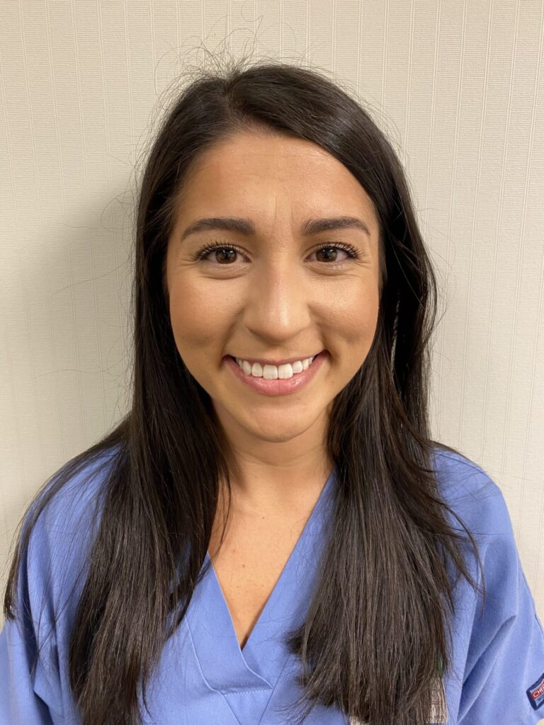 A woman with long dark hair, wearing blue medical scrubs, smiles at the camera while standing in front of a cream-colored, textured wall.