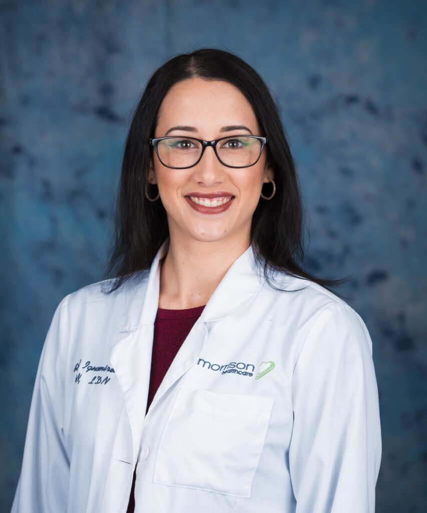 A woman with long dark hair and glasses, wearing a white lab coat with Morrison Healthcare and embroidered text, smiles while standing in front of a blue mottled background.