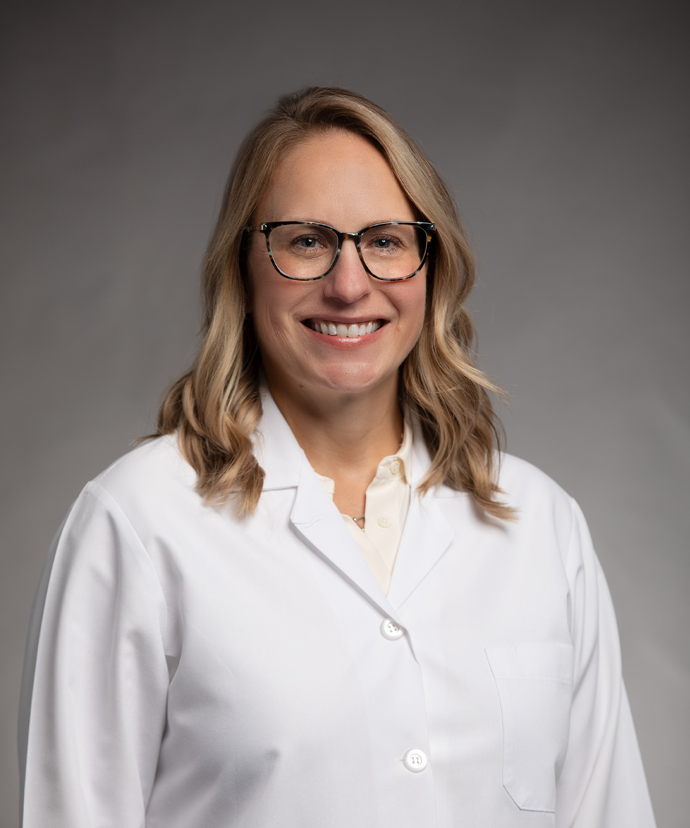 A smiling woman with blonde hair and glasses wears a white lab coat and stands against a plain gray background, representing the compassionate care at Fort Sanders Perinatal Center.