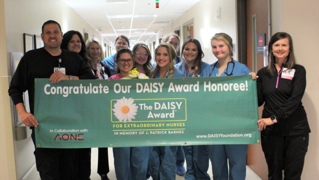 A group of nurses and healthcare staff smile in a hospital hallway, proudly holding a green DAISY Award banner that reads, Congratulate Our DAISY Award Honoree! The DAISY Award for Extraordinary Nurses.
