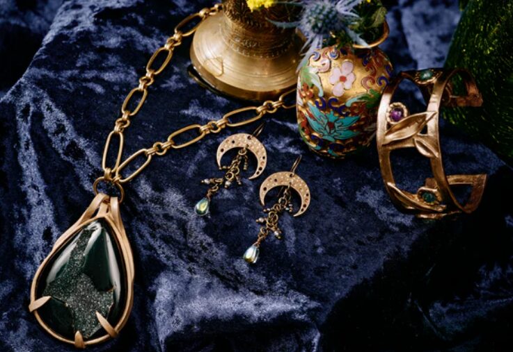 A collection of vintage-style jewelry on dark blue velvet by Tony Perrin, featuring a large green stone pendant on a gold chain, crescent moon earrings, a gold bracelet with gems, and a decorative vase.