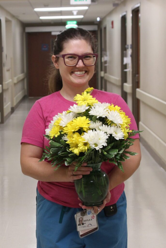 A smiling person with glasses, wearing a pink shirt, blue pants, and an ID badge, holds a bouquet of yellow and white flowers in a green vase—celebrating their DAISY Award—while standing in a hallway.