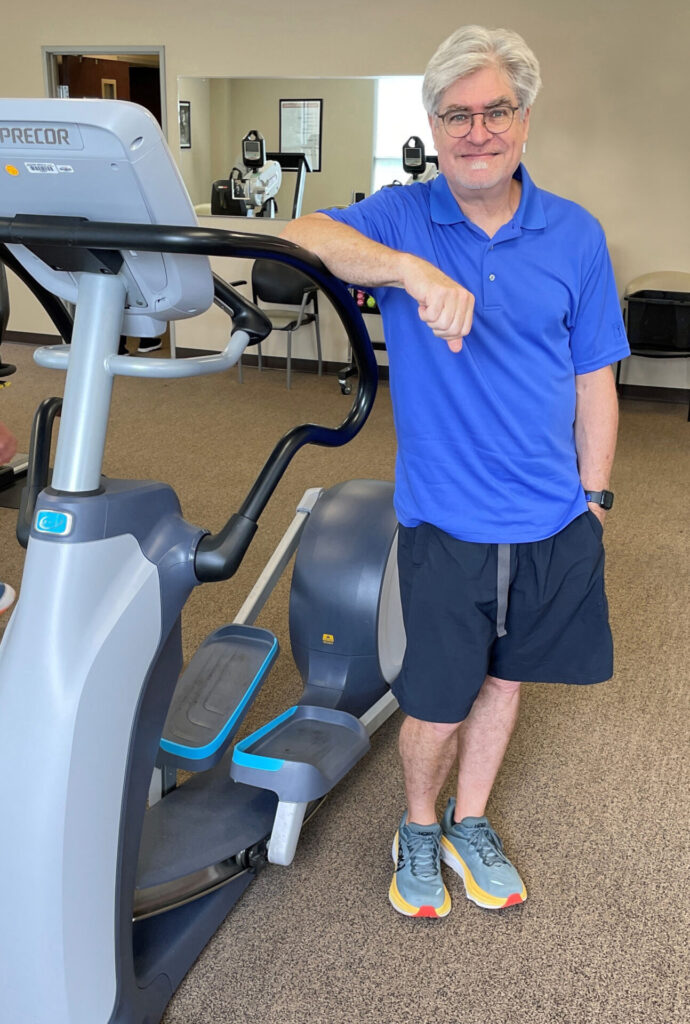 John Hook in blue t-shirt standing by exercise bike.