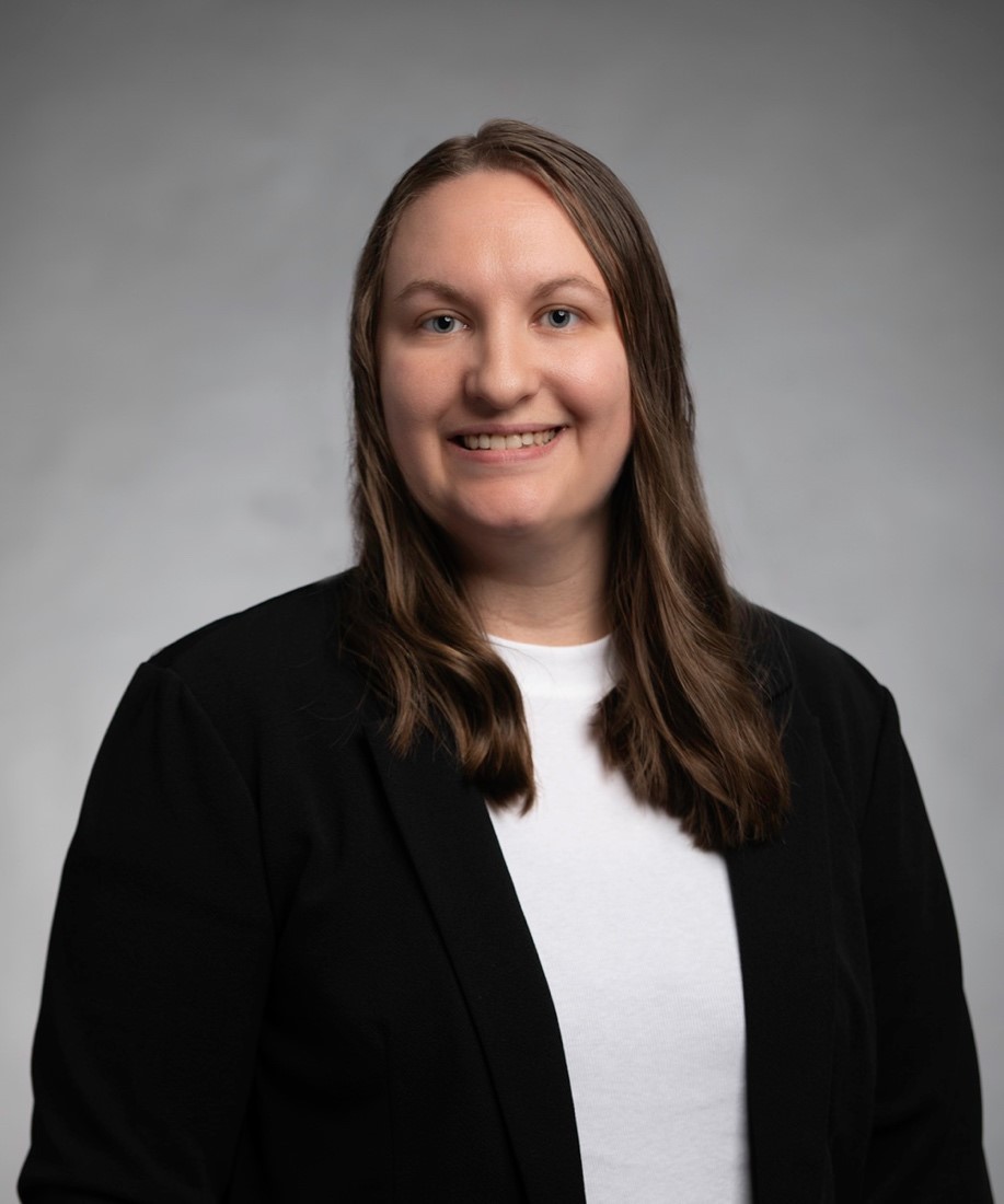 A woman with long brown hair wearing a black blazer over a white shirt, smiling at the camera, posed against a plain gray background—confident and ready for new opportunities in cancer careers.