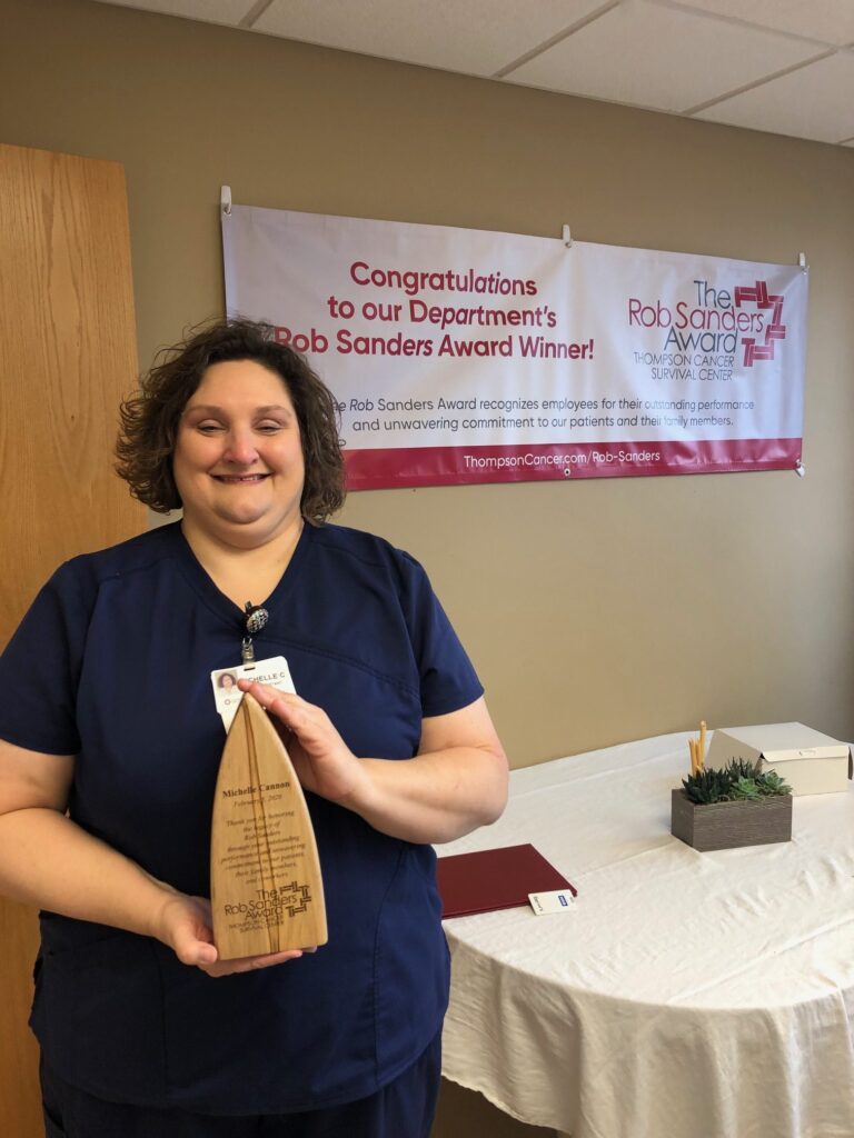 A woman in navy scrubs stands indoors, smiling and holding a trophy. Behind her is a banner that reads, Congratulations to our Department’s Rob Sanders Award Winner!” with a table and decor nearby.