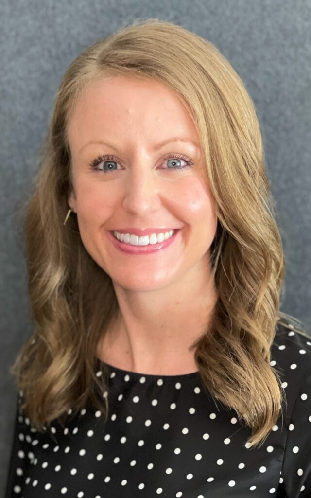 Kasey Smith, Pharm D 5 6 22 A woman with wavy, shoulder-length blonde hair smiles at the camera, radiating positivity. She wears a black top with white polka dots and stands in front of a plain gray background, embodying hope often seen in those undergoing cancer treatments.