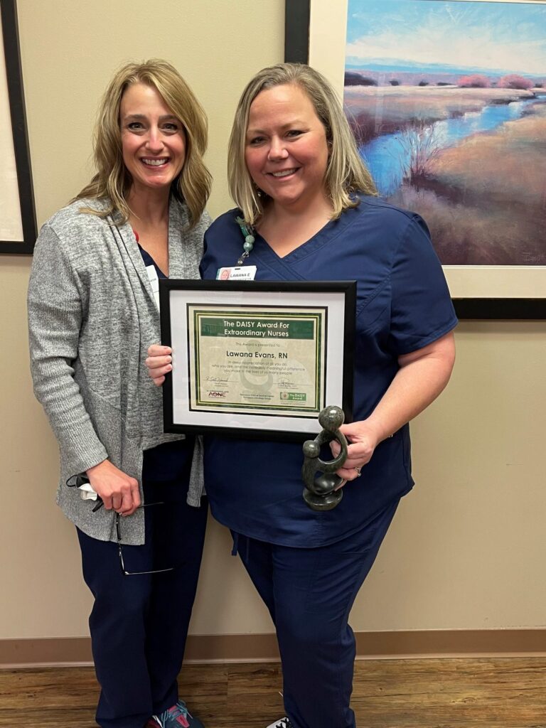 Two women in navy blue scrubs, both DAISY nurses, smile in an office. One holds a framed DAISY Award for Extraordinary Nurses certificate and a statuette. A scenic landscape painting hangs on the wall behind them.