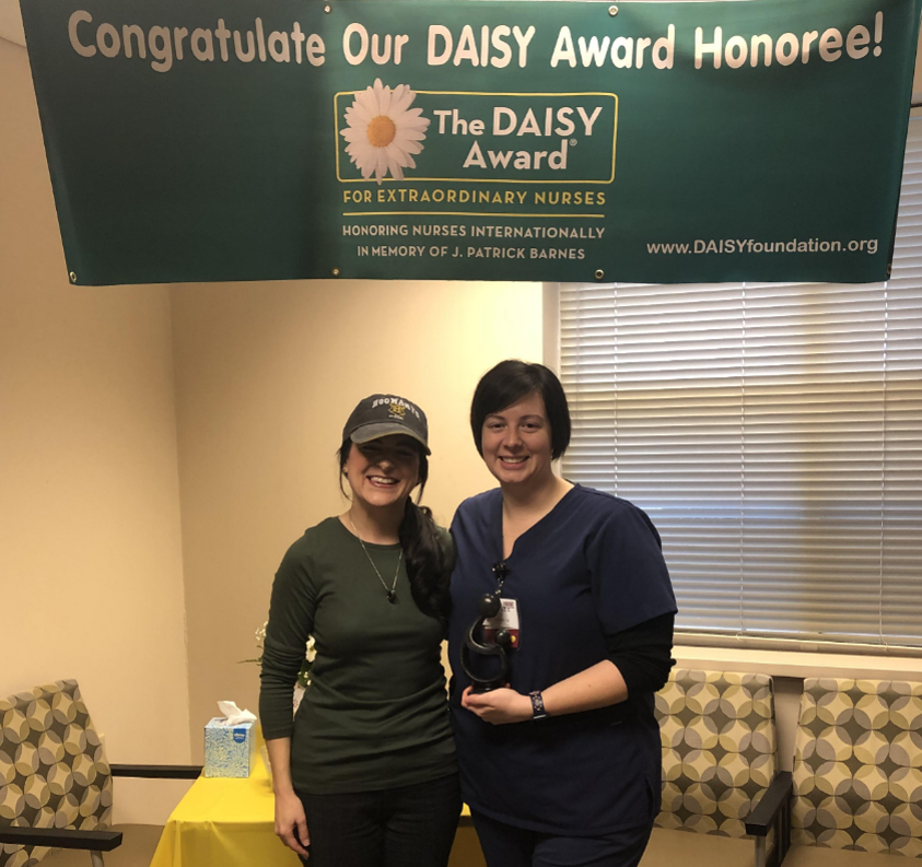 Two women standing and smiling under a banner that reads Congratulate Our DAISY Award Honoree! One woman is in scrubs holding an award, while the other wears casual clothes and a hat. They are in a room with chairs and a table.