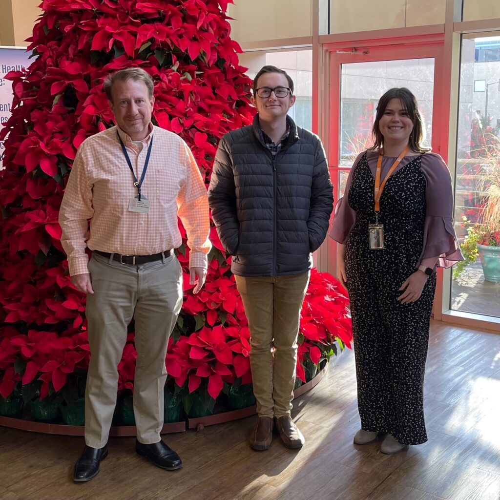 Three people stand smiling in front of a large, decorated poinsettia tree inside a sunlit room. The two men and one woman, professionals in medical physics, are wearing work badges and casual business attire.