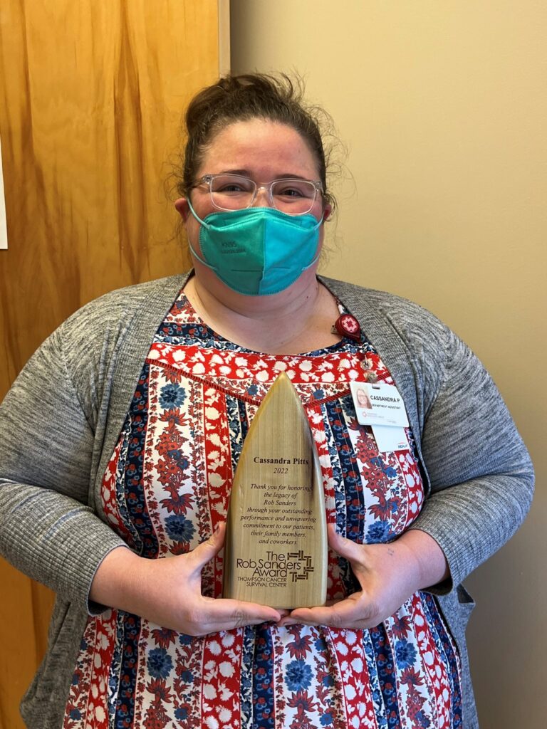 A person wearing a teal face mask, glasses, and a patterned dress holds a wooden award. They are standing indoors against a beige wall and wooden door. A name badge is clipped to their cardigan.