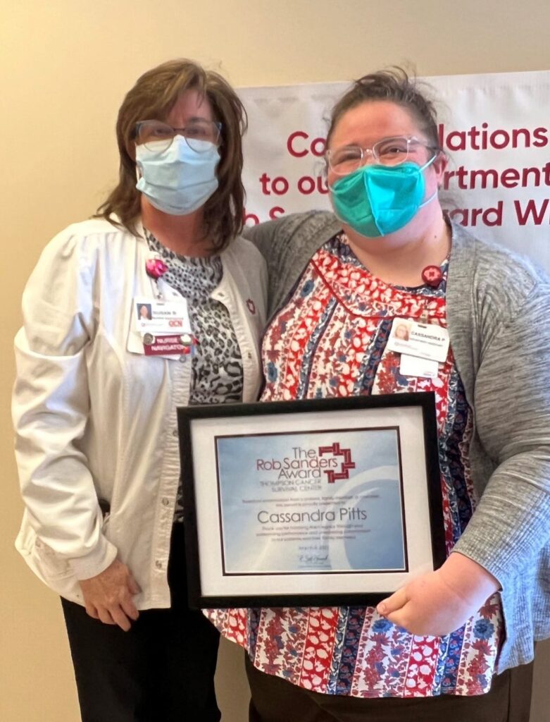 Two women wearing face masks stand together. One woman is holding a framed certificate reading The Rob Sanders Award with the name Cassandra Pitts. Both are smiling and dressed in work attire, standing in front of a congratulatory banner.