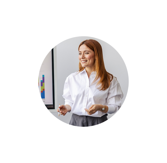 A woman with long red hair, wearing a white blouse and gray skirt, stands smiling and gesturing in front of a chart with colorful bars on a whiteboard, presenting data from Covenant Health LLD courses.