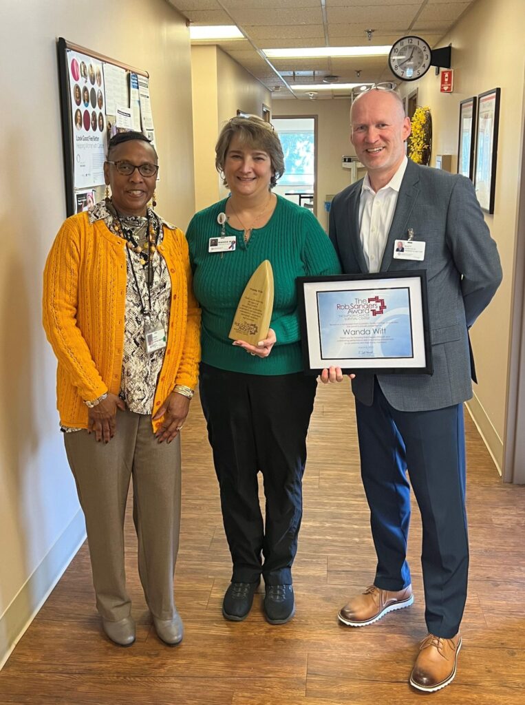 Three people stand in a hallway; the woman in the center holds a trophy, and the man on the right holds a framed certificate. They are smiling and appear to be celebrating an achievement.