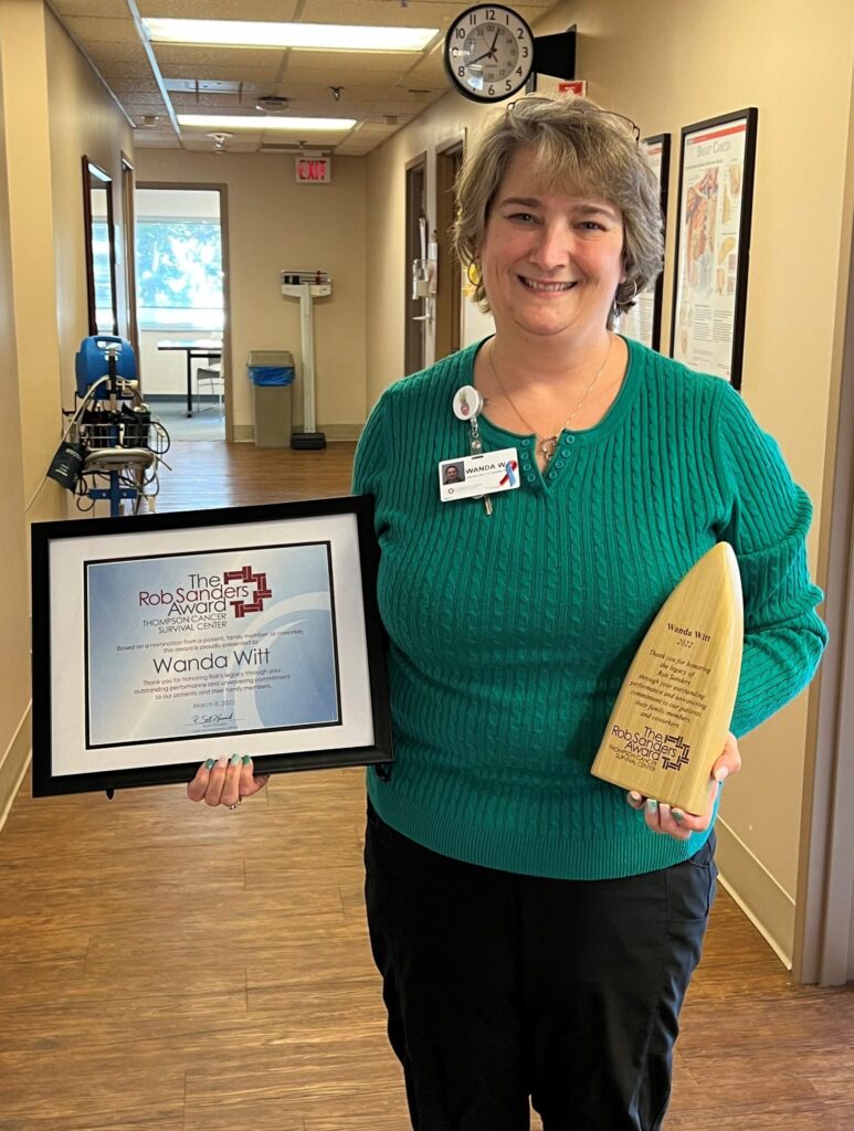 A smiling woman in a green sweater stands indoors, holding a framed certificate and a wooden award. She appears to be in a medical or office setting, with visible equipment and posters in the background.