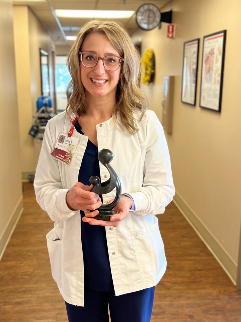 A woman in a white lab coat stands in a hallway, smiling and proudly holding a small abstract DAISY Award trophy. She has shoulder-length blond hair, glasses, a name badge, and is framed by charts and a clock on the wall behind her.