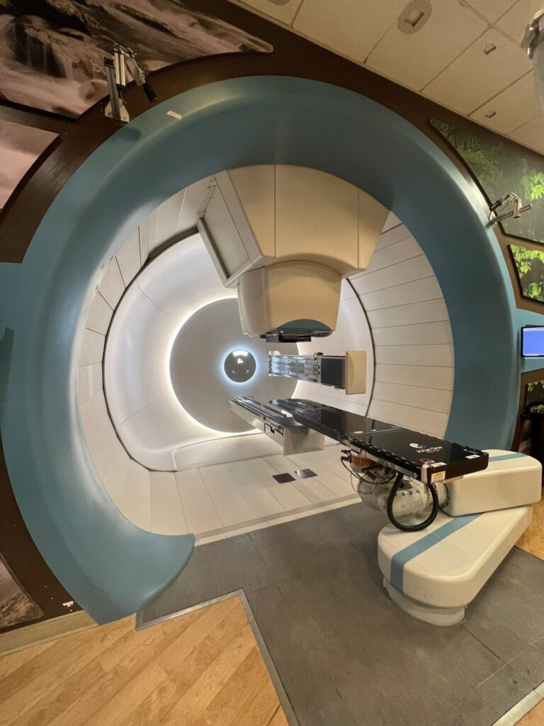 A modern medical machine for radiation therapy, as seen on the Thompson Proton Center tour, features a large circular gantry, patient table, and control panels in a clinical setting with wood and white décor.