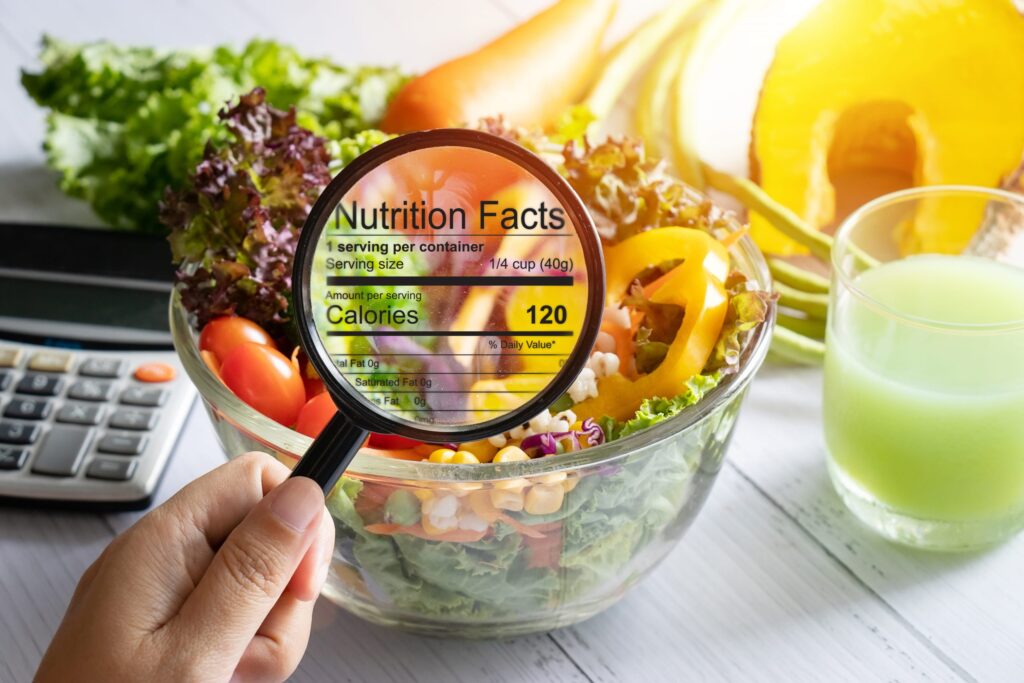 A hand holds a magnifying glass over a salad bowl, enlarging the nutrition facts label showing 120 calories per serving. A calculator, green juice, and fresh vegetables are displayed on a white table in the background.