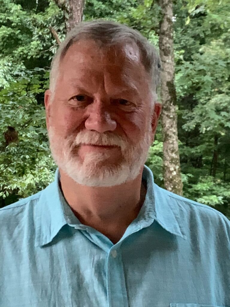 An older man with short gray hair and a white beard, wearing a light blue collared shirt, smiles at the camera after shoulder surgery while standing outdoors in front of green, leafy trees.